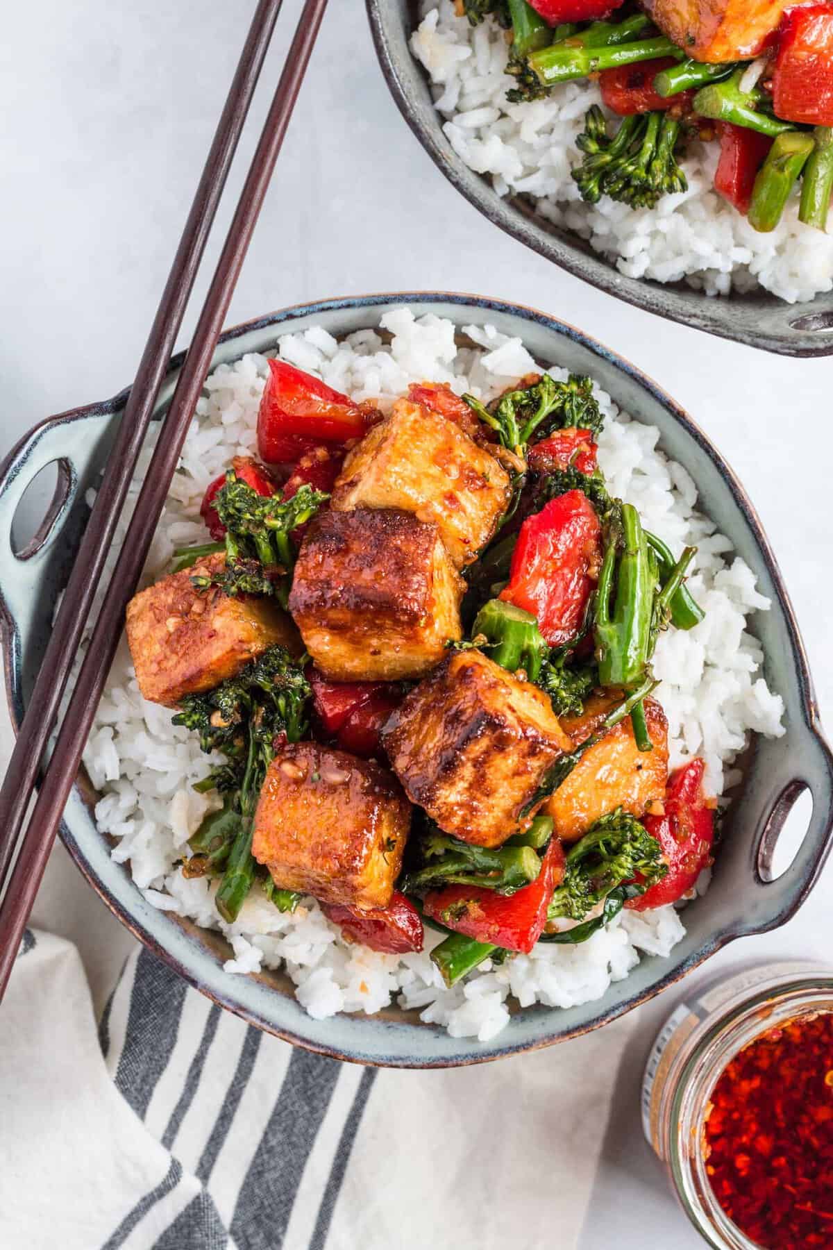 Two bowls of white rice and tofu stir fry are placed on a white surface and are ready to be eaten. 