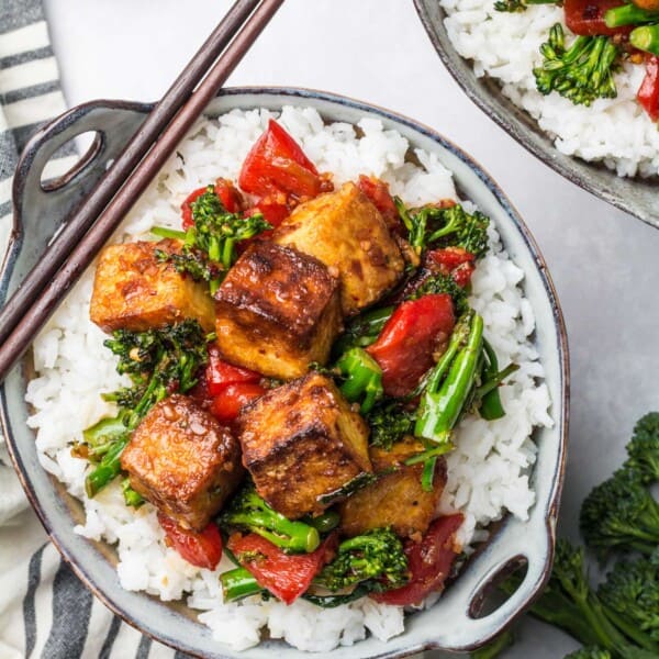A serving of spicy tofu stir fry sits on a bed of white rice in a small bowl. Chopsticks sit on the side of the bowl.