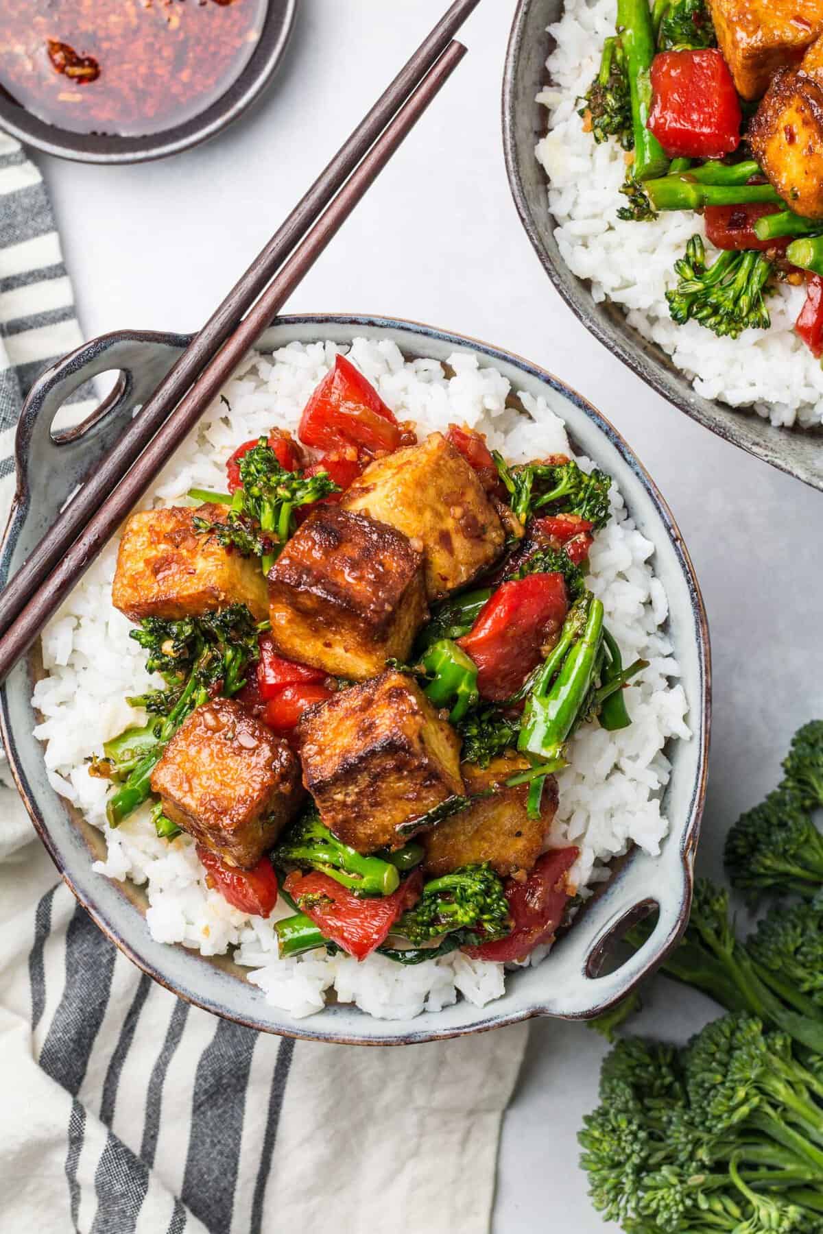 A serving of spicy tofu stir fry sits on a bed of white rice in a small bowl. Chopsticks sit on the side of the bowl.