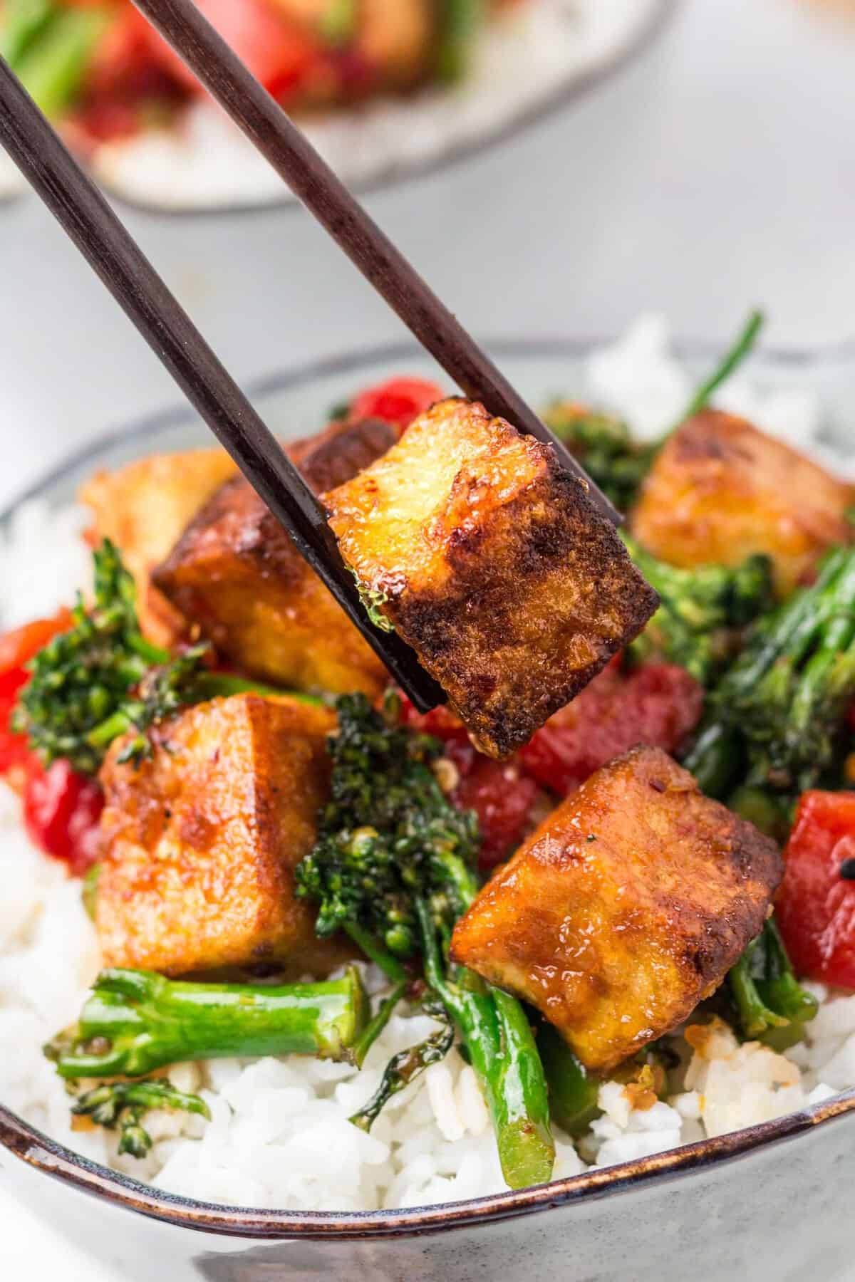A piece of crispy tofu is being lifted from the bowl by a pair of dark brown chopsticks. 