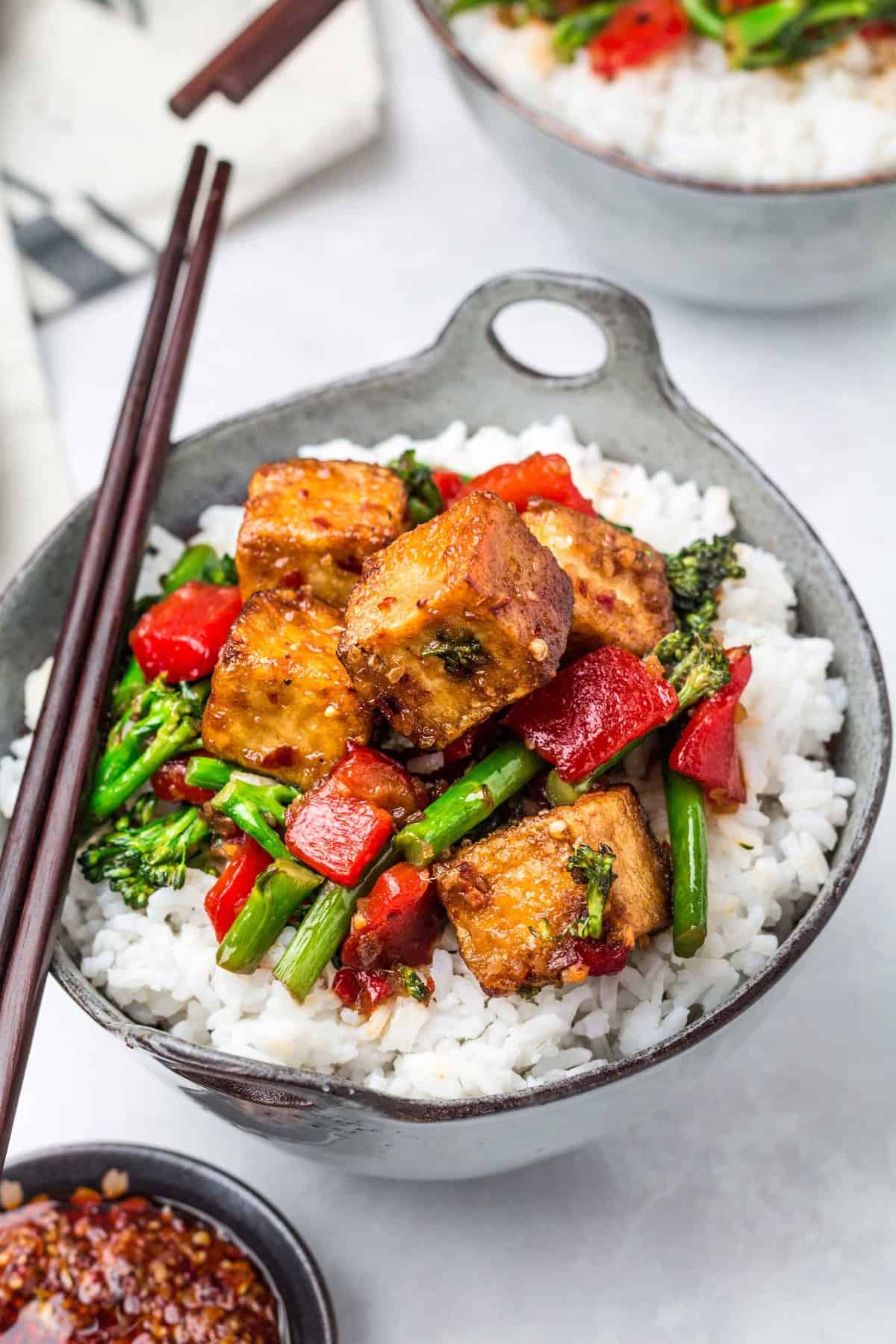 A bowl of rice and stir fry sits next to a small bowl of chili crunch sauce on a white countertop. 