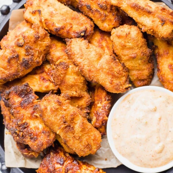 A number of crispy fish sticks are on a large serving platter with a small bowl of creamy cajun dipping sauce.