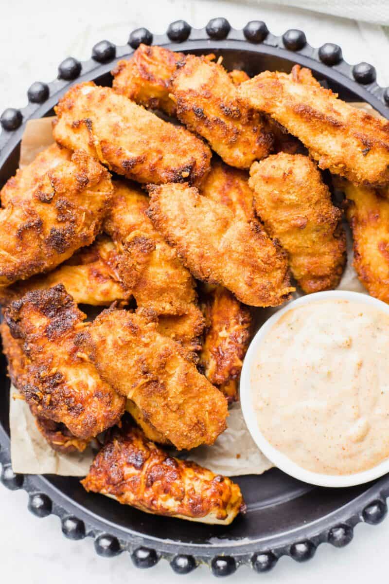 A number of crispy fish sticks are on a large serving platter with a small bowl of creamy cajun dipping sauce.