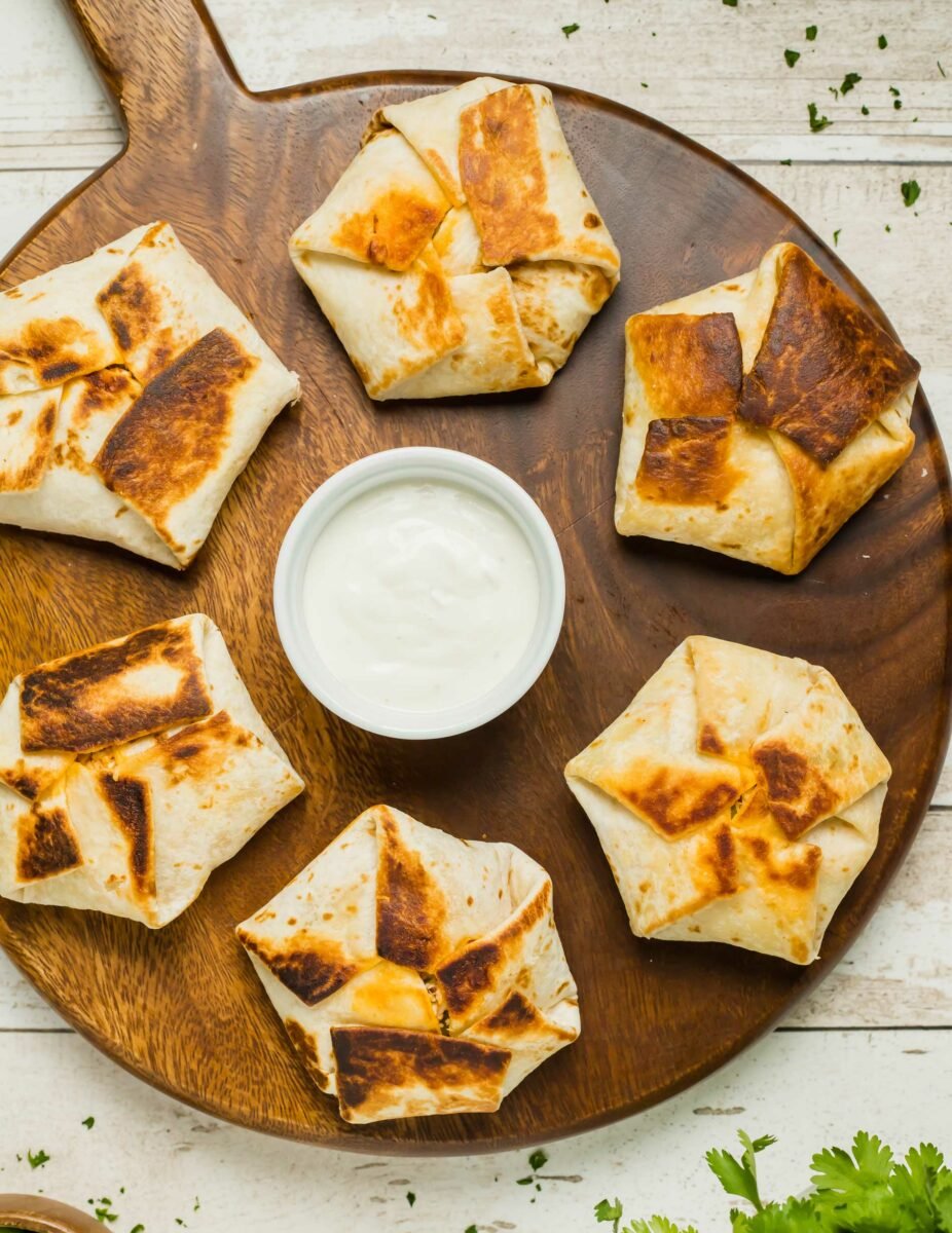 Six mini buffalo chicken crunchwraps are placed on a wooden serving platted with a small white bowl of dip in the middle.