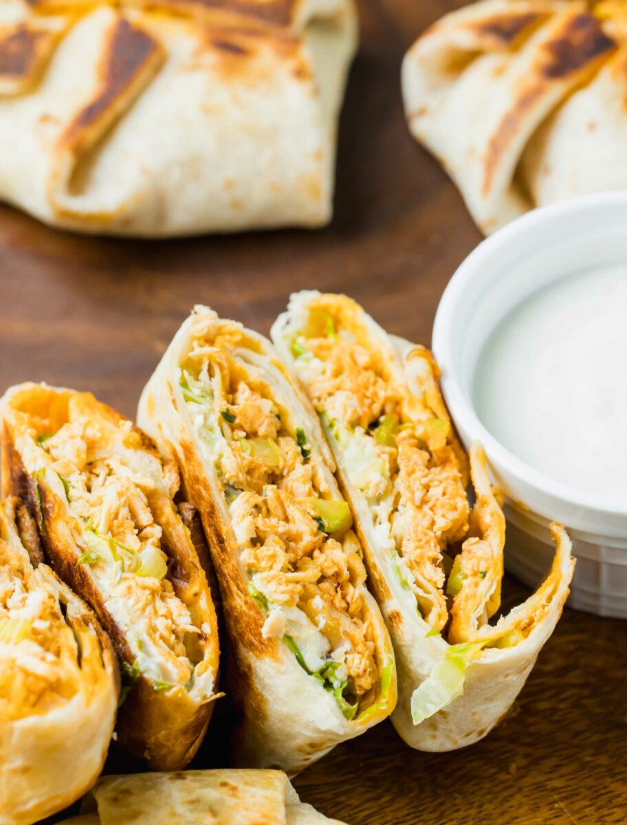 Mini chicken crunchwraps have been sliced in half and are placed on a brown surface next to a small white bowl of dip. 