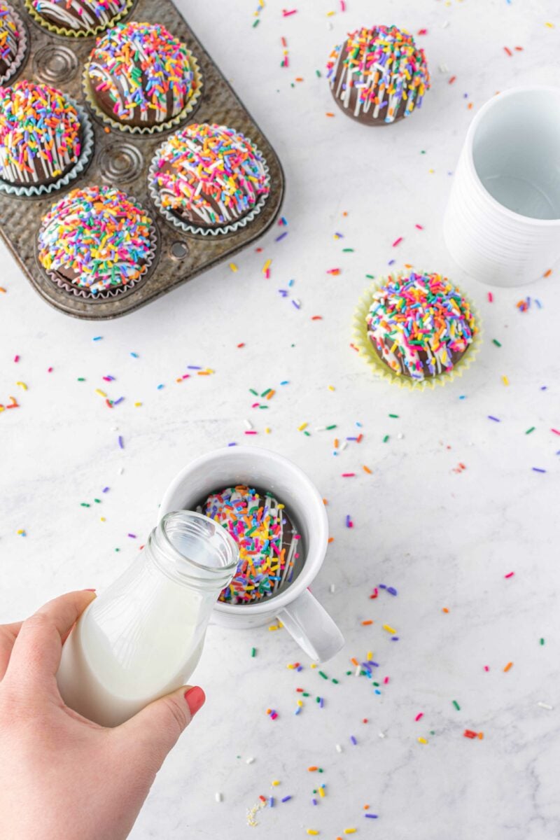 Milk from a jaw is being poured onto a funfetti cocoa bomb in a white mug.