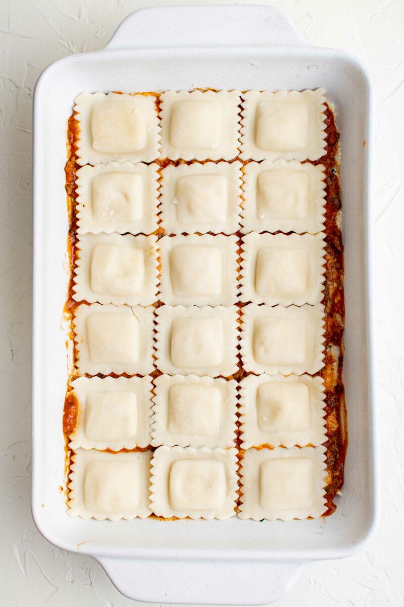 A layer of cooked ravioli is placed on top of red sauce in a baking dish.