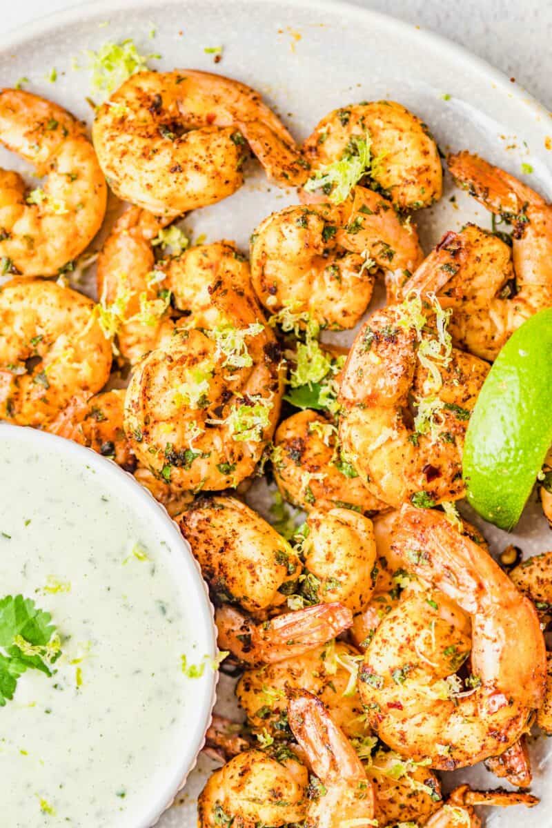 A large plate of shrimp is being served next to a bowl of cilantro lime dressing. 