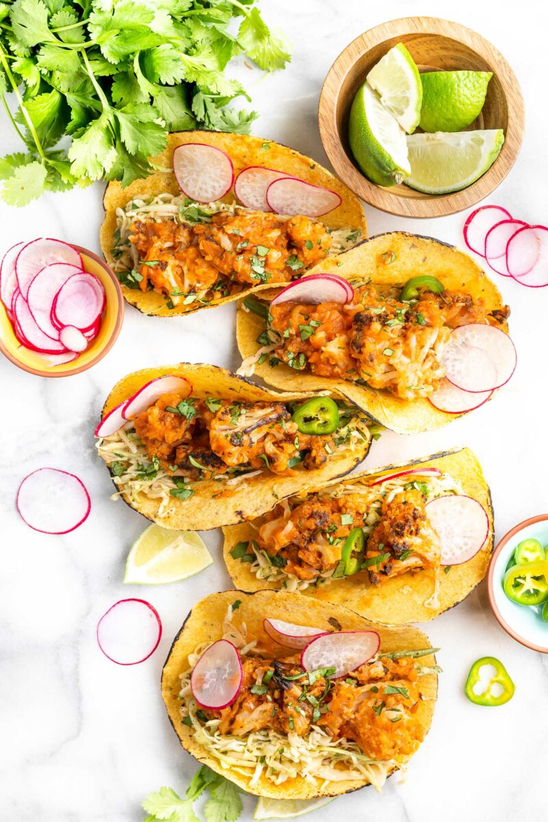 Five cauliflower tinga tacos are placed on a white surface.