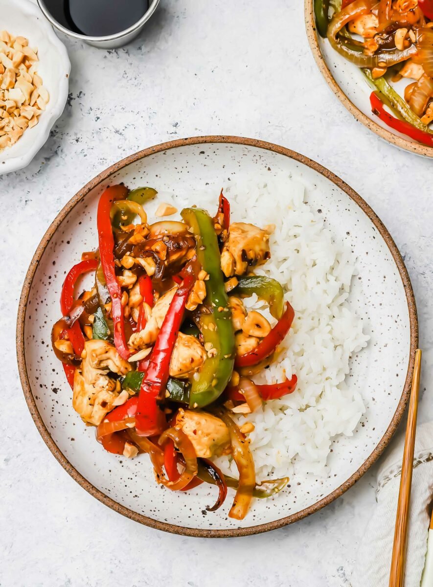 A serving of Kung Pao chicken is placed next to cooked white rice on a white spotted plate. 
