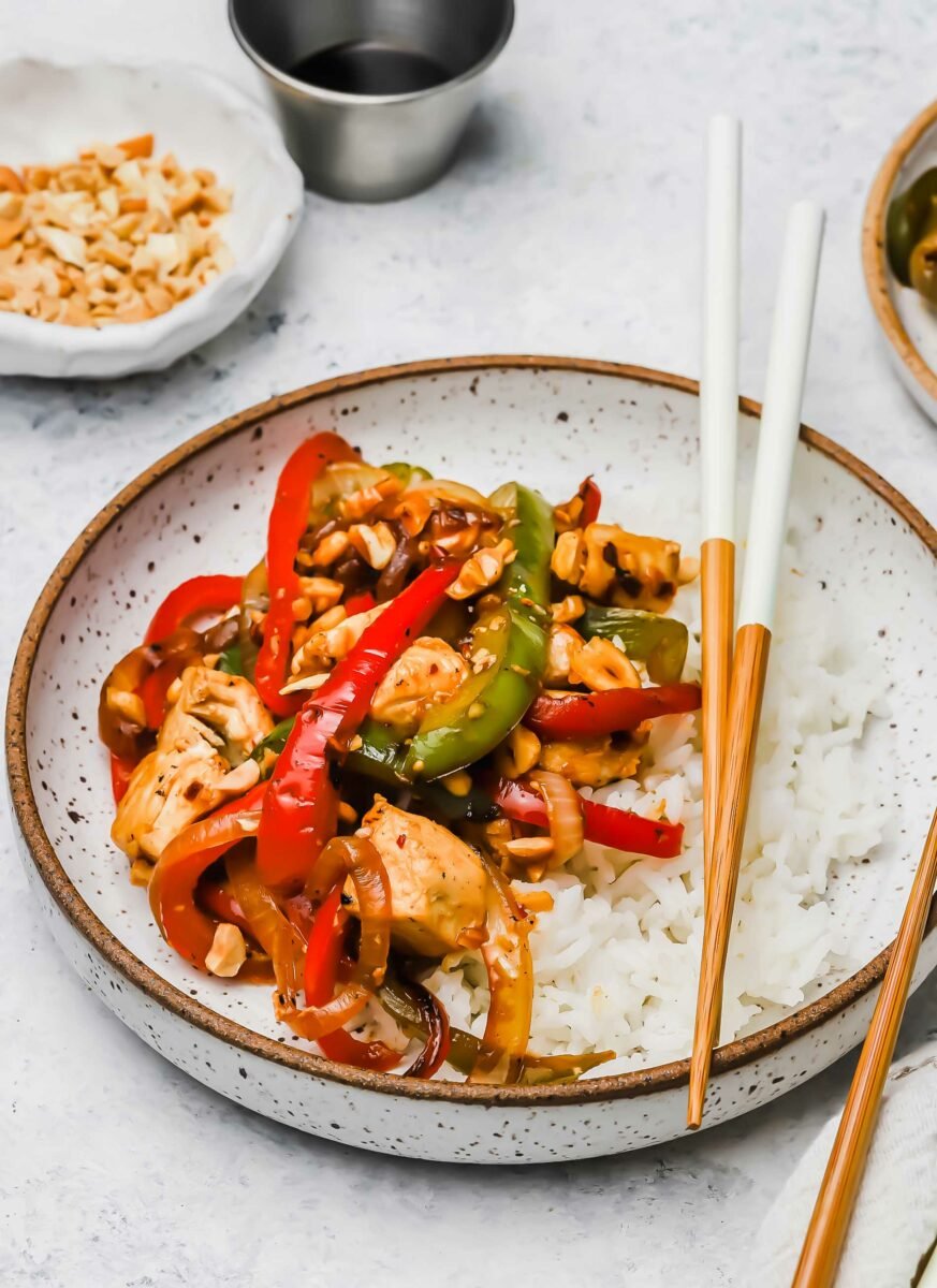 Chopsticks sit on a white speckled bowl filled with rice and Kung Pao chicken. 