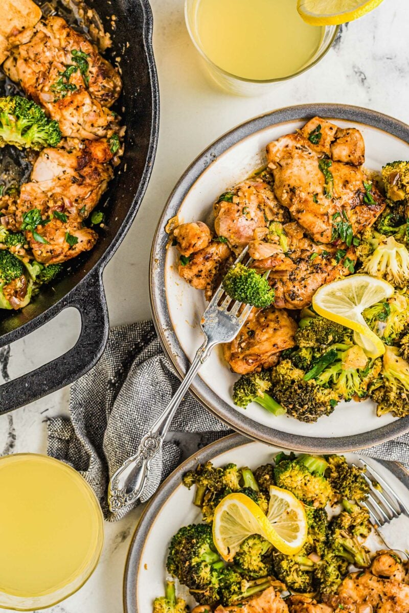 Two white plated with servings of lemon garlic chicken and broccoli are placed next to a skillet. 