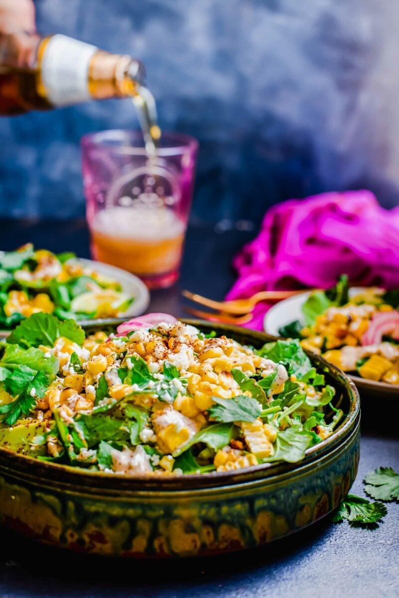 A beer is being poured in the background of a Mexican corn and kale salad. 