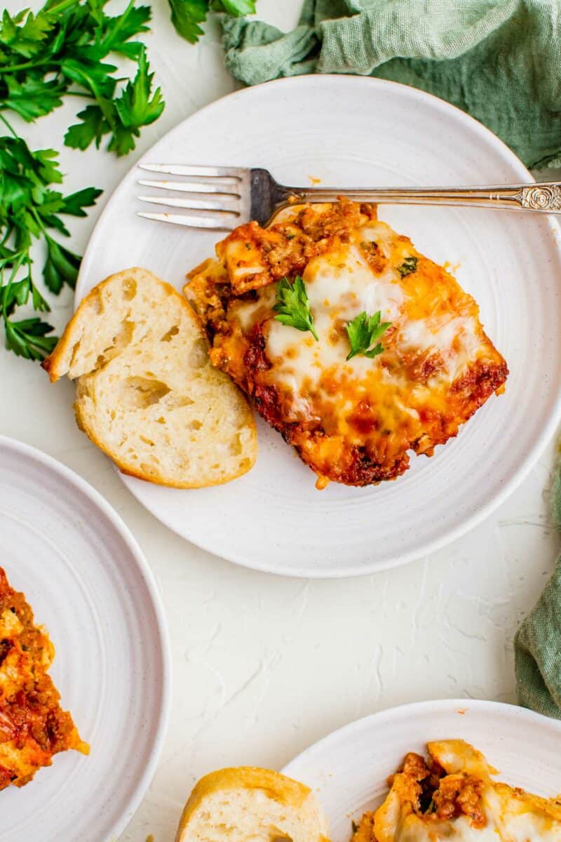 A piece of lasagna is on a white plate with a piece of bread next to it.