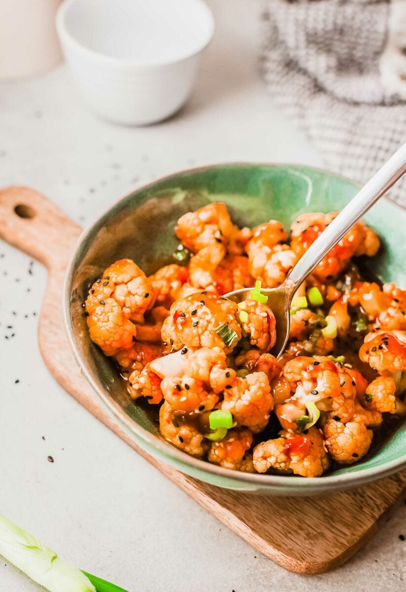 A small spoon is picking up a few pieces of sticky Korean-style cauliflower out of a green bowl.