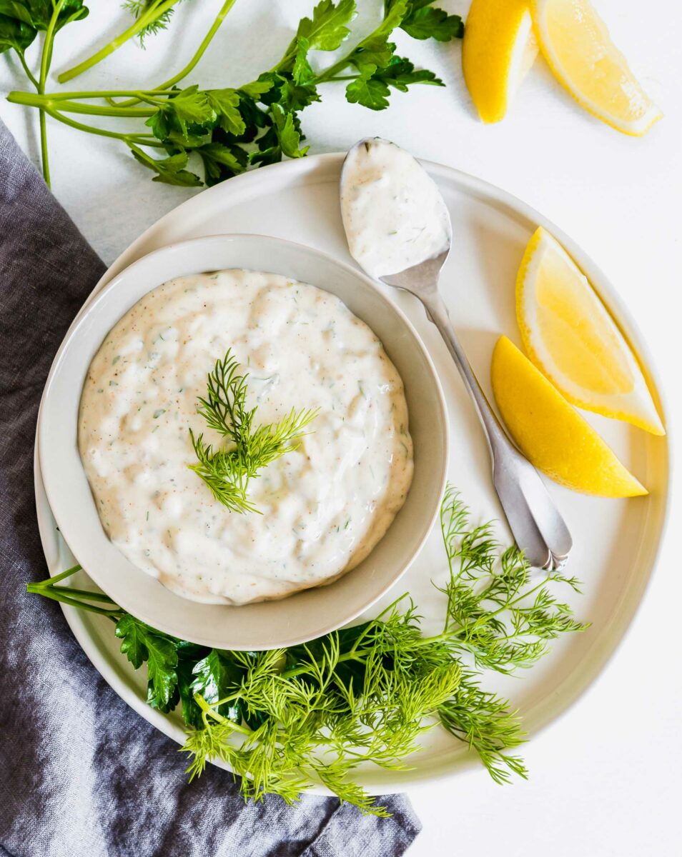 A bowl full of tartar sauce in on a plate next to dill, lemon slices, and a spoon.
