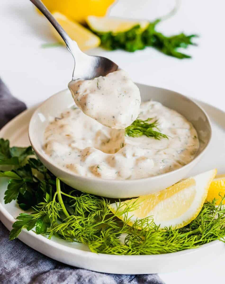 A metal spoon is lifting a bite sized portion of tartar sauce out of the full bowl.