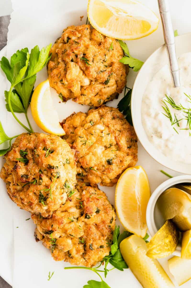 Four crab cakes are presented on a white plate with fresh herbs. 