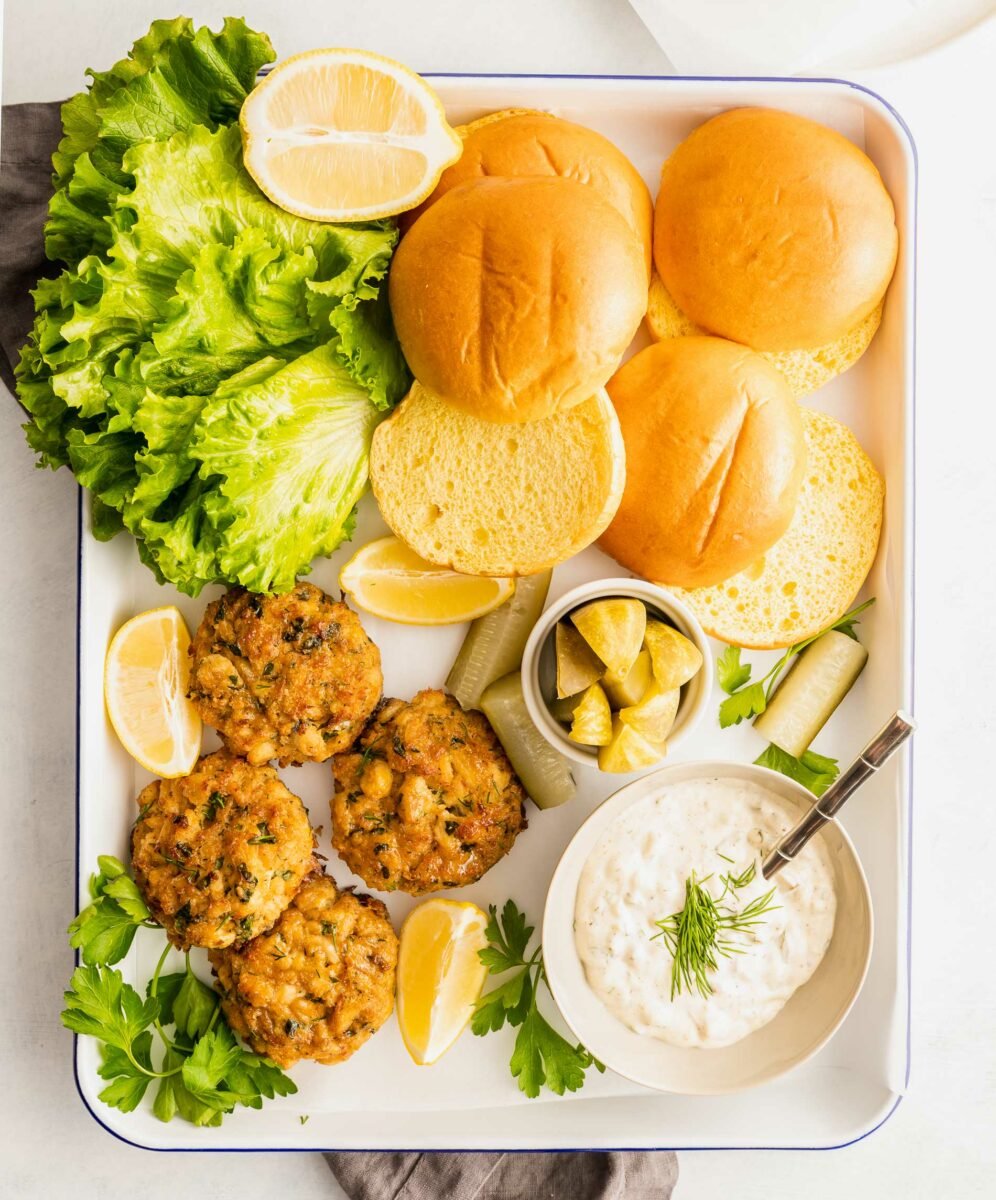 Crab cakes, buns, lettuce and sauce are on a white serving platter. 