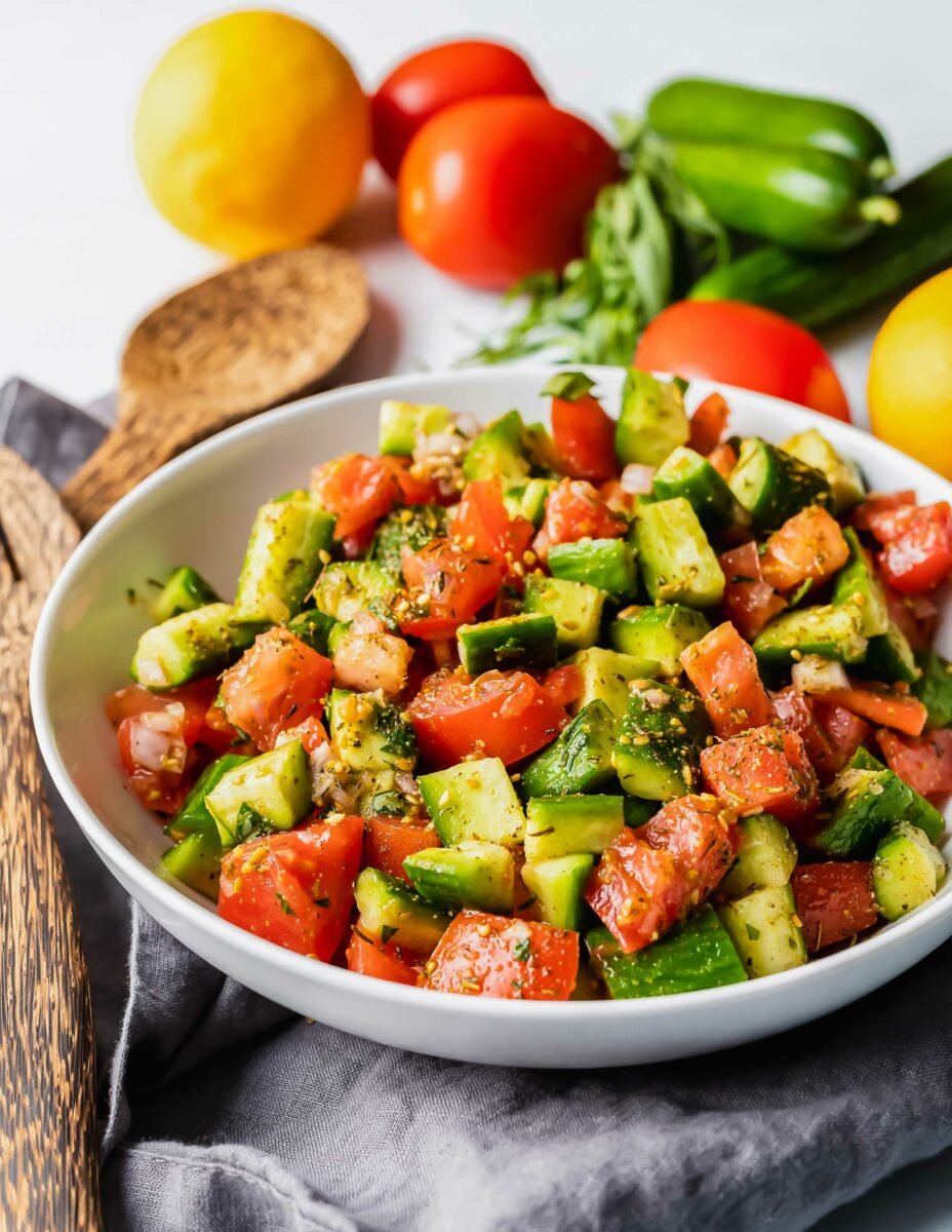 A tomato and cucumber salad has been tossed with wooden salad tossers next to uncut vegetables. 