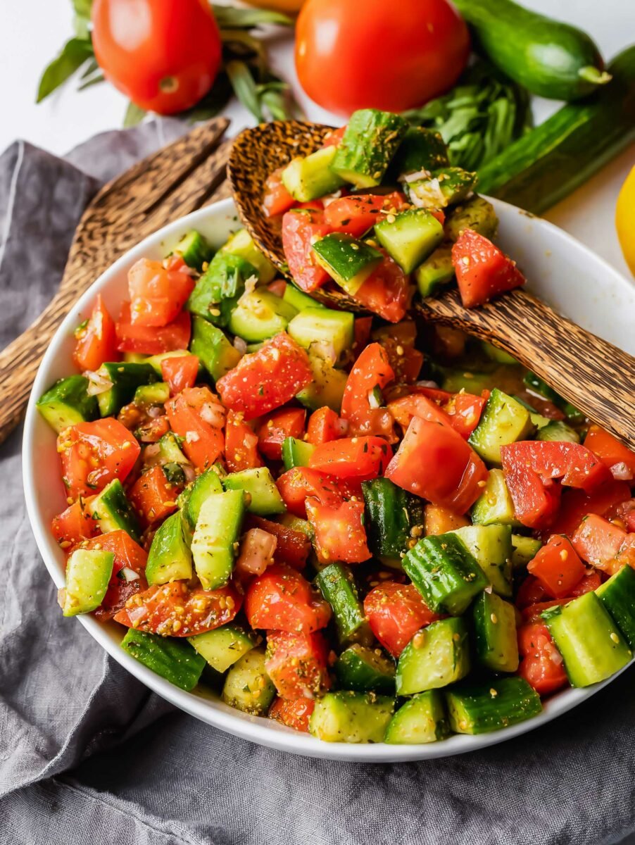 A large wooden spoon is lifting a small serving of tomatoes and cucumbers from the bowl. 