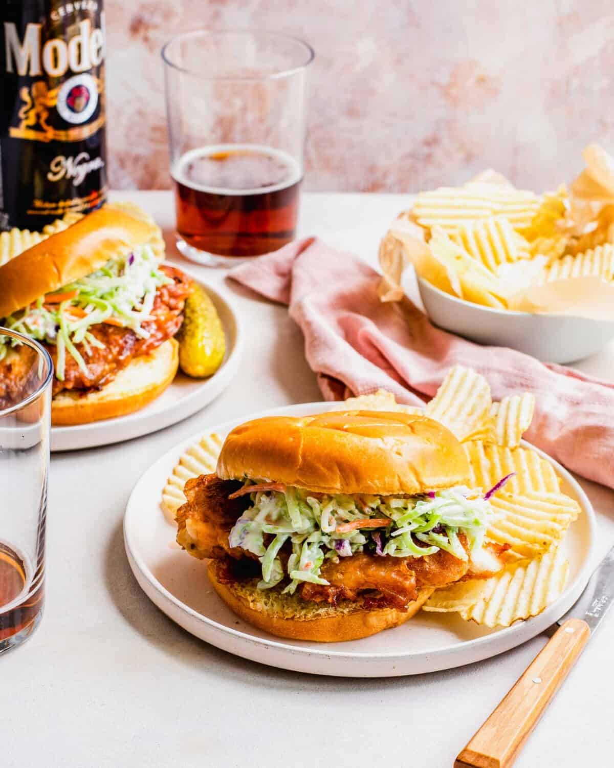 Two plates with chicken sandwiches are placed next to a cup of beer and extra potato chips.