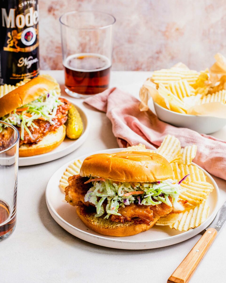 Two plates with chicken sandwiches are placed next to a cup of beer and extra potato chips. 