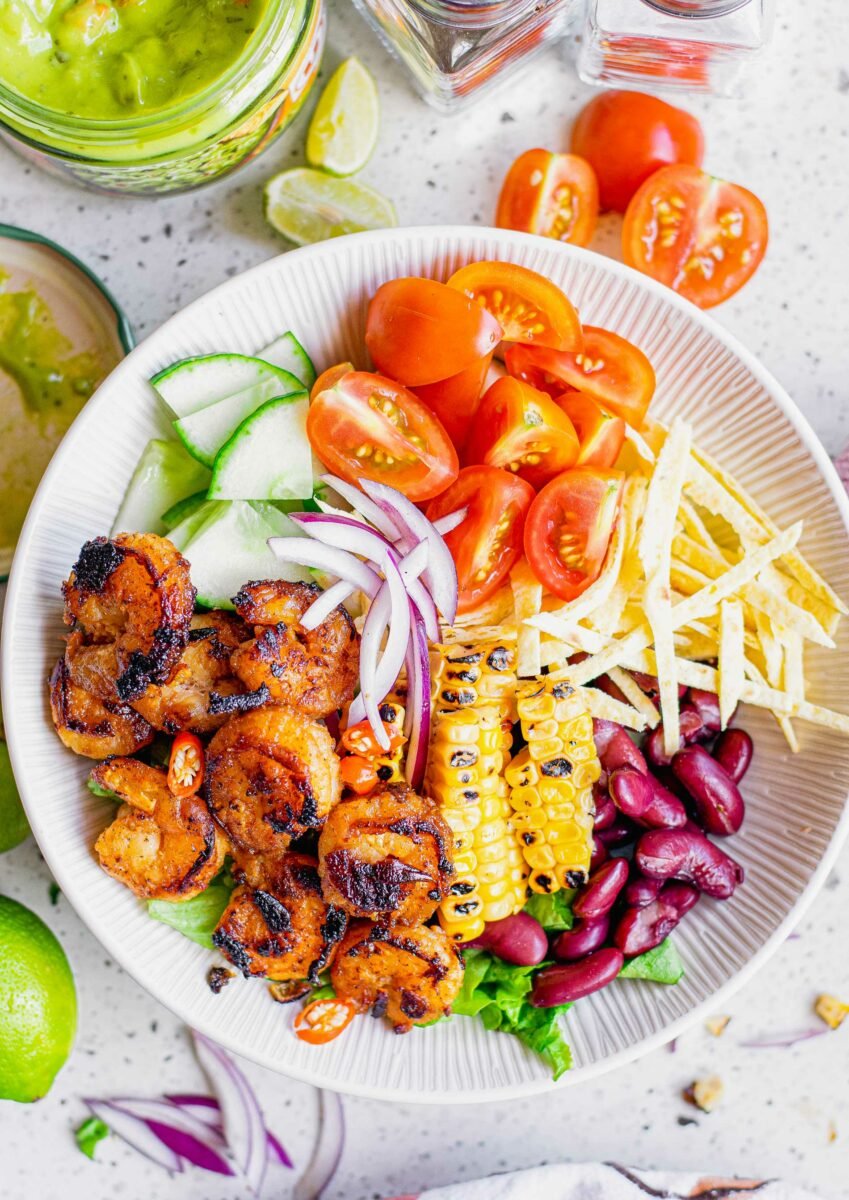 A white bowl is filled with cucumbers, onions, tomatoes, tortilla strips, corn, kidney beans, and shrimp. 