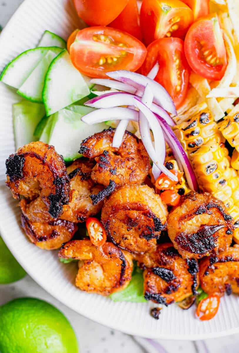 Blackened shrimp are placed next to onions, corn and cucumbers in a white bowl. 