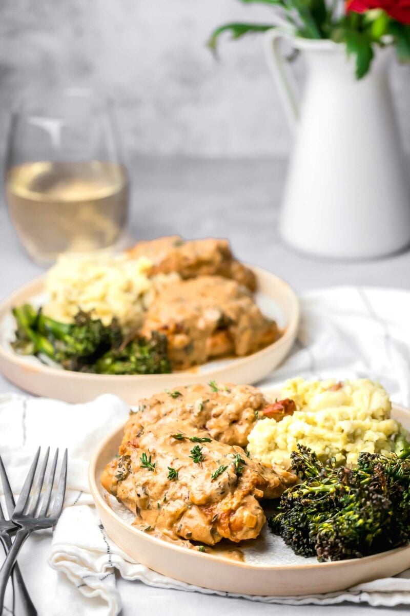 A plate with chicken thighs, broccolini and mashed potatoes is placed on a white tea cloth. 