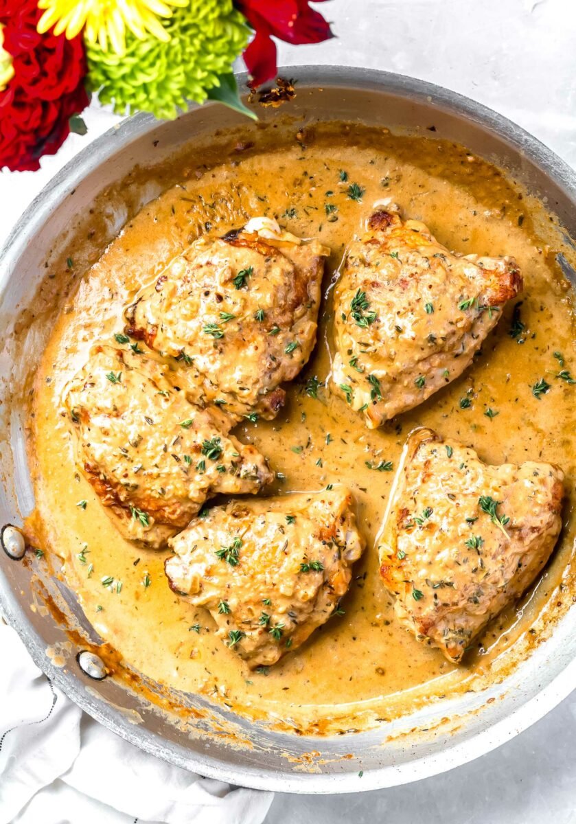 Five chicken thighs are in a skillet filled with creamy yellow mustard sauce. 