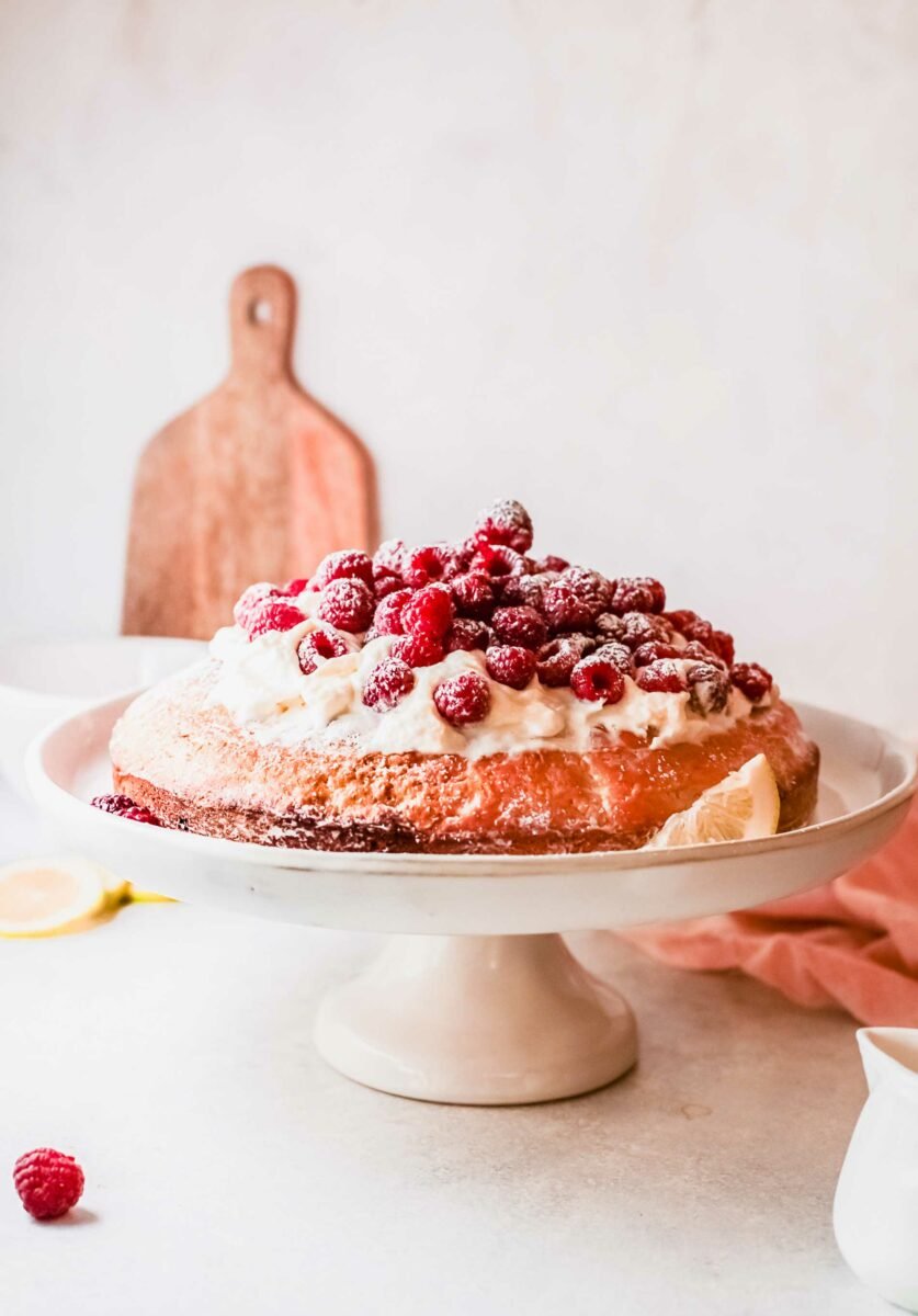 A raspberry olive oil cake is on a white cake stand. 