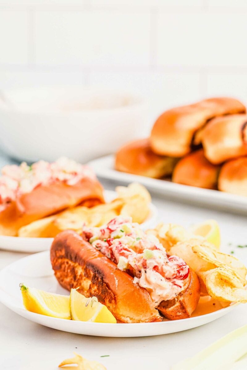 Lemon wedges, a lobster roll, and chips are on a white plate. 