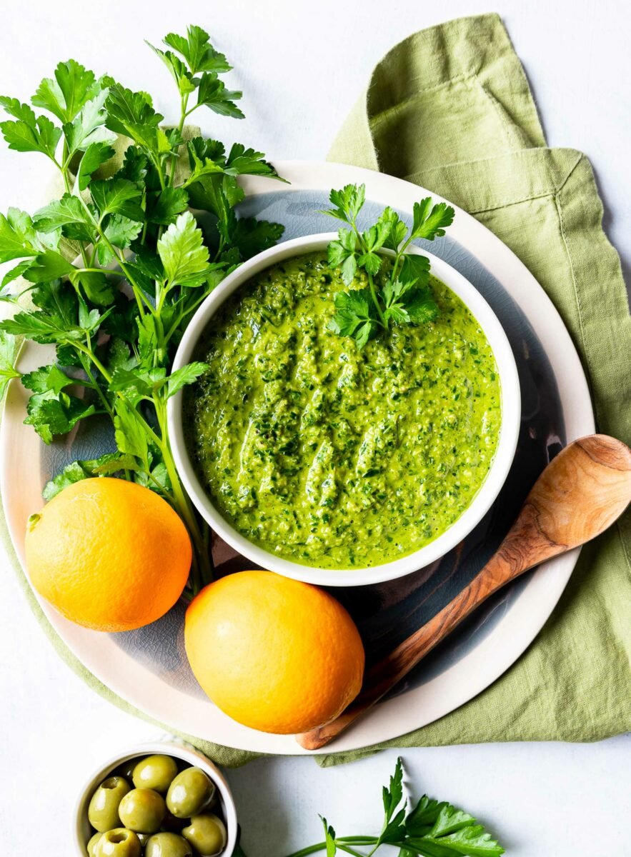 Herbs and lemons are placed next to a white bowl filled with gremolata. 