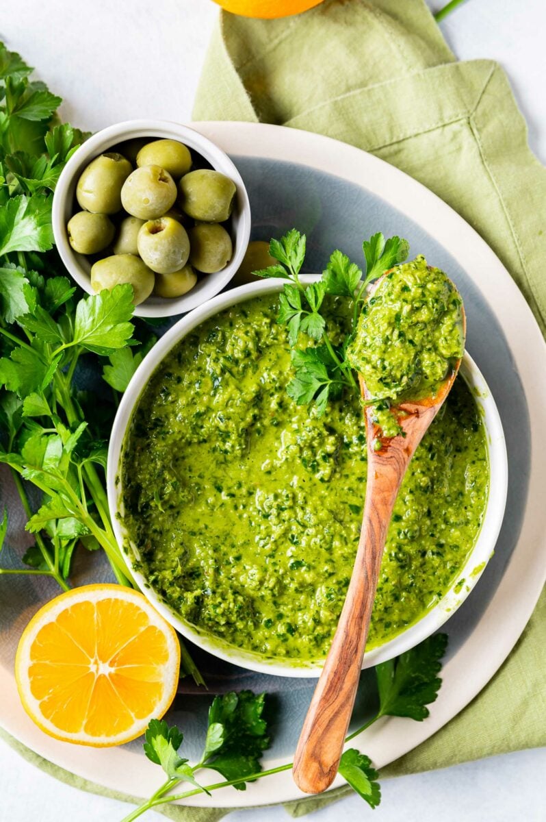 Gremolata is in a bowl next to lemons, herbs, and a wooden spoon. 