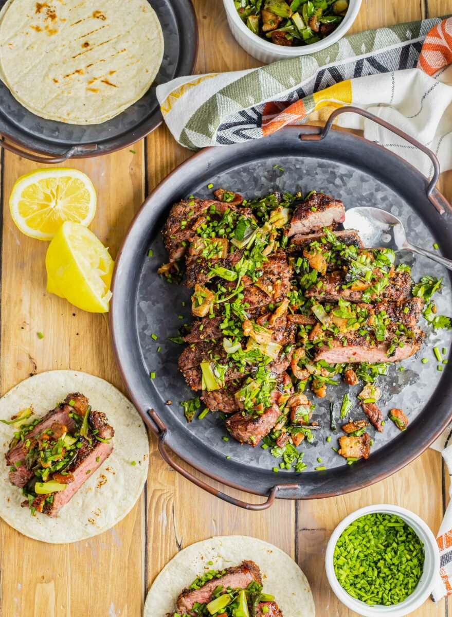 Squeezed lemons and filled corn tortillas are placed next to a large portion of steak and herb topping. 