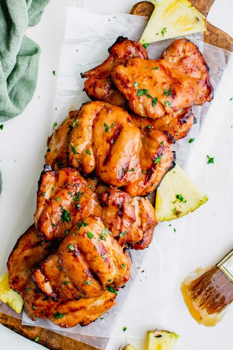 Multiple grilled chicken thighs are placed on a sheet of parchment paper with small pieces of pineapple. 