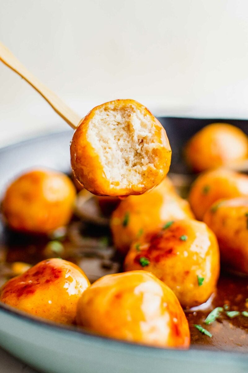 A skewer is lifting a bitten into meatball from the pan. 