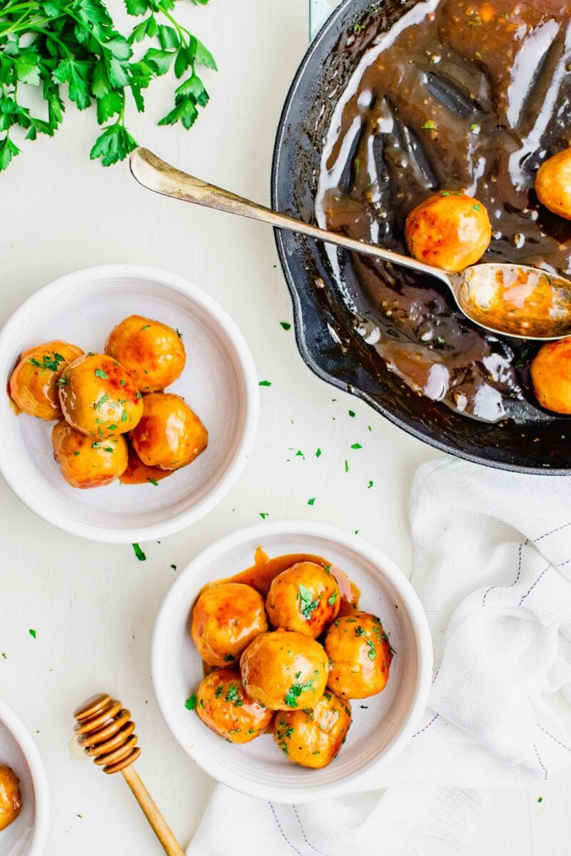 Two white bowls with meatballs are placed next to a black skillet with meatballs. 