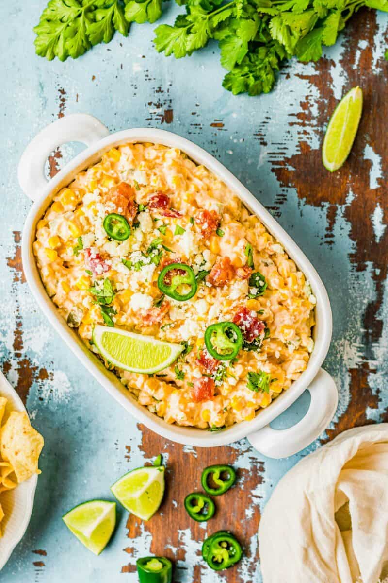 Sliced limes and jalapenos are placed on top of a white container filled with corn dip. 