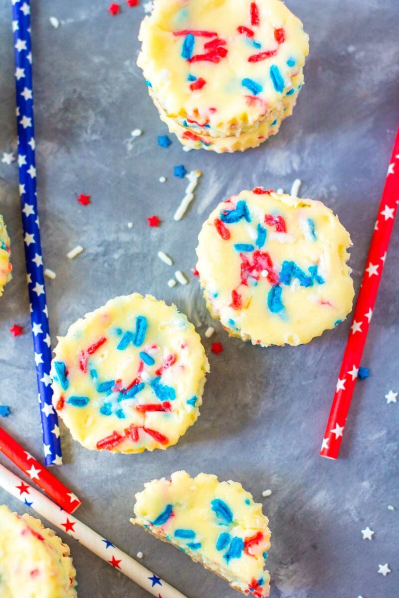 Several mini cheesecakes are placed on a gray surface with sprinkles surrounding them. 