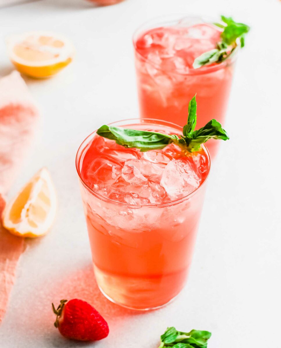 Two strawberry gin rickeys are placed on a white counter top and garnished with basil. 