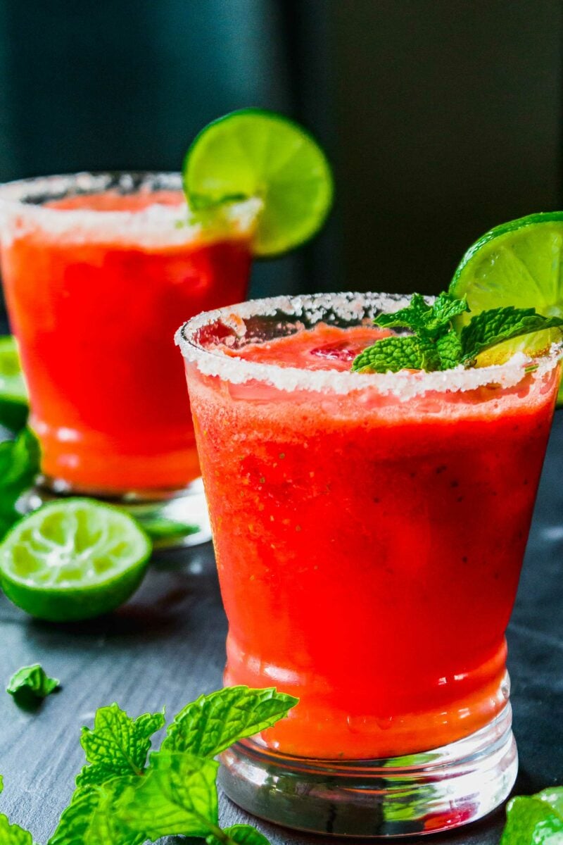 Mint leaves and limes are placed around two cups on a dark surface. 