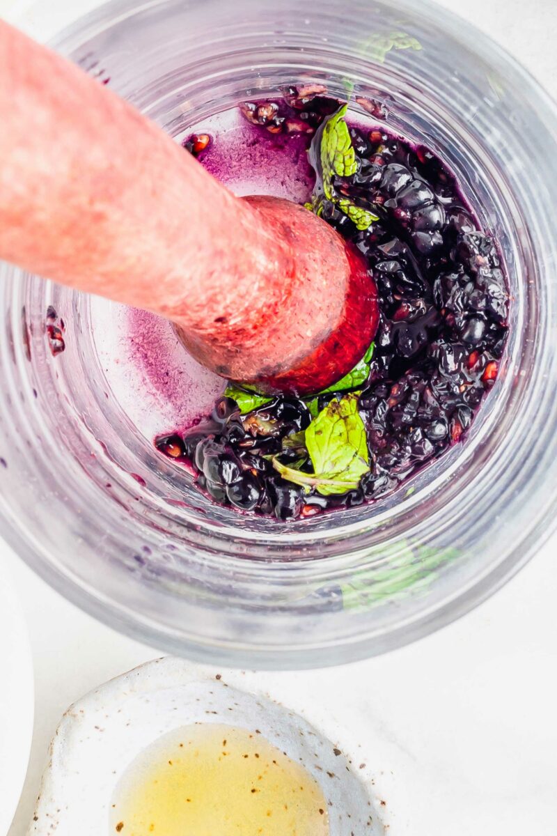 A muddler is smashing berries and mint at the bottom of a glass. 