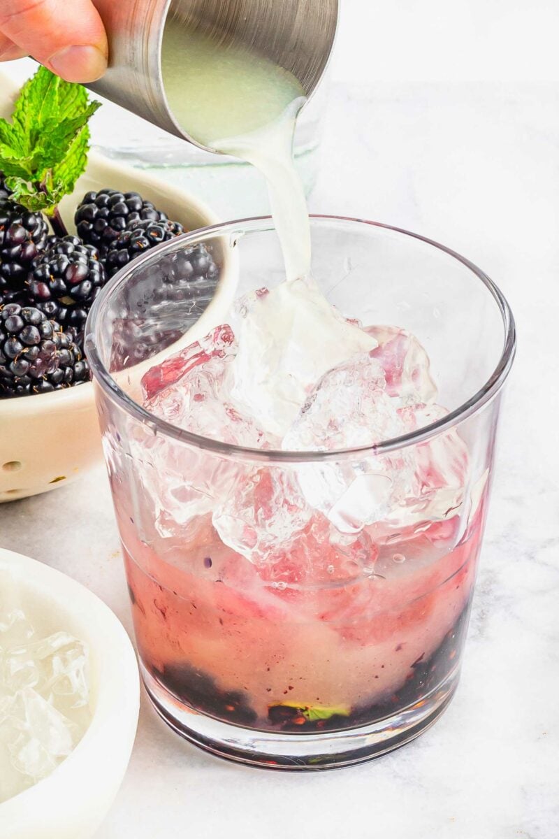 Lime juice is being poured into a glass with muddled berries and mint at the bottom of it.