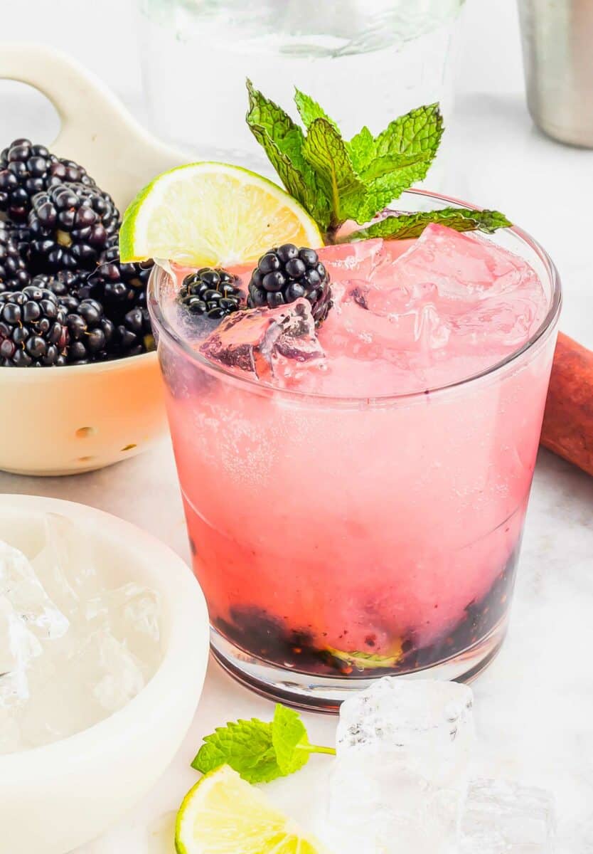 A glass is filled with a pink mojito and is garnished with berries, lime, and mint. 