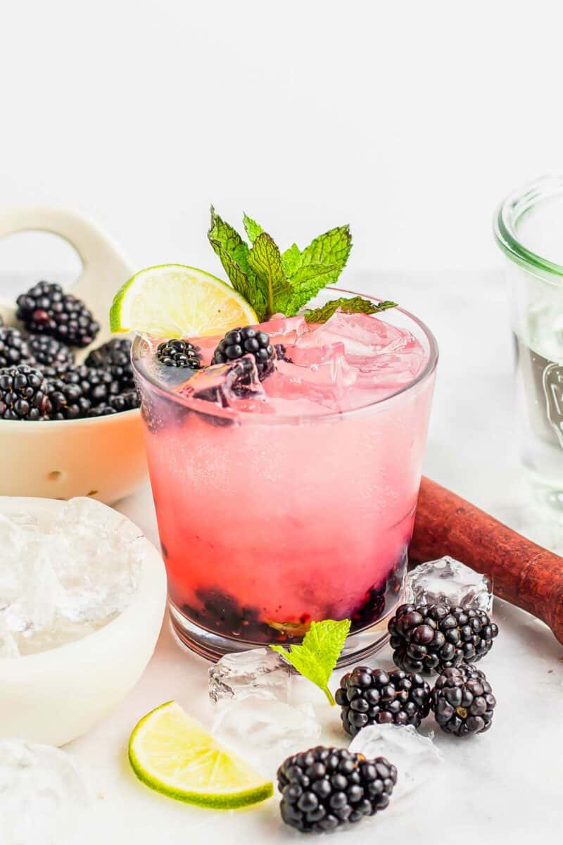 Several blackberries and ice cubes are placed near a full glass of mojito. 