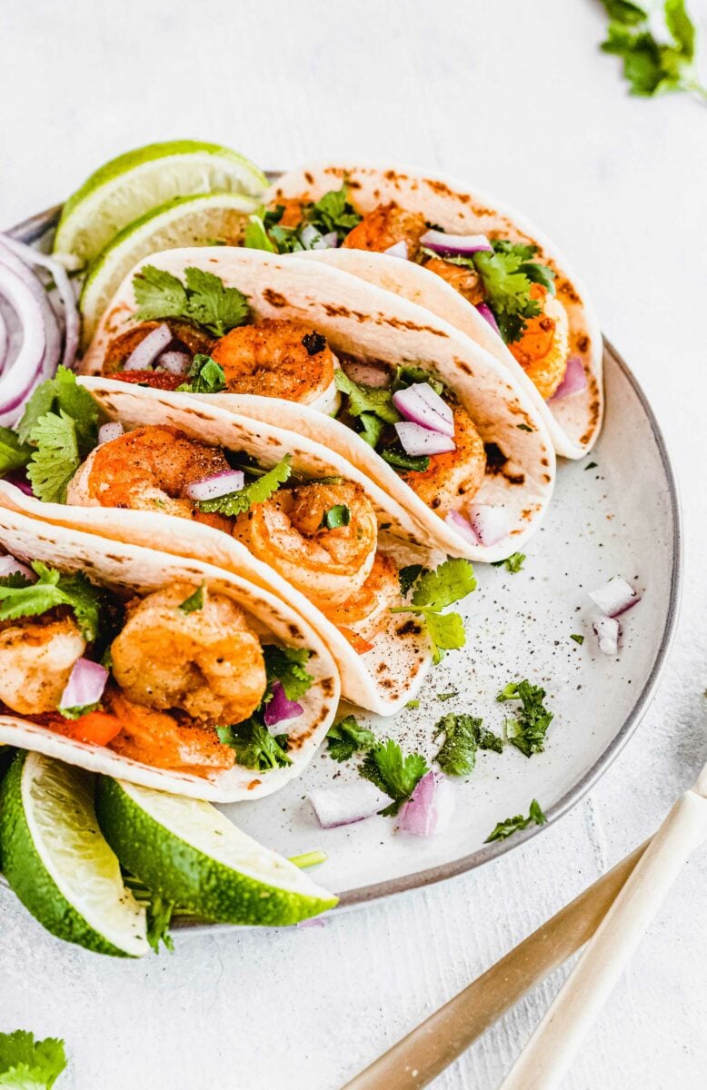 Several tacos are placed on a white plate with fresh cilantro and lime wedges. 