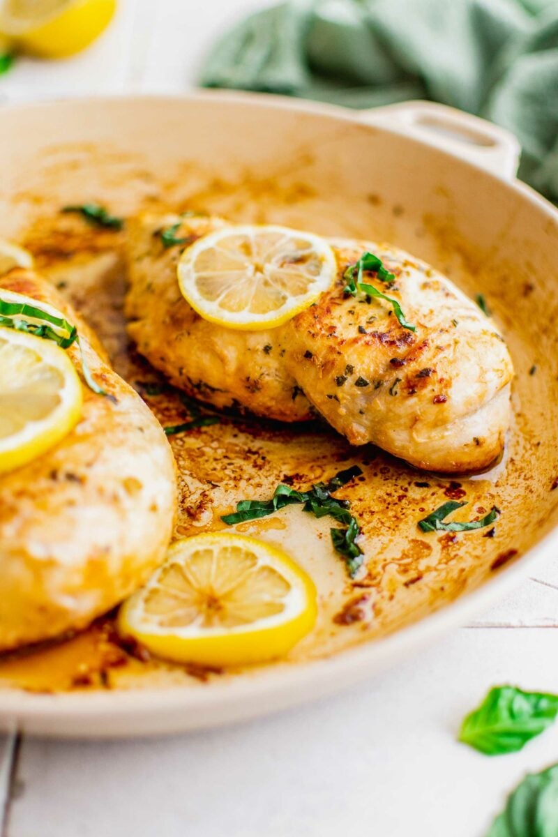 Two chicken breasts are cooking in a pan with lemon, olive oil and basil. 