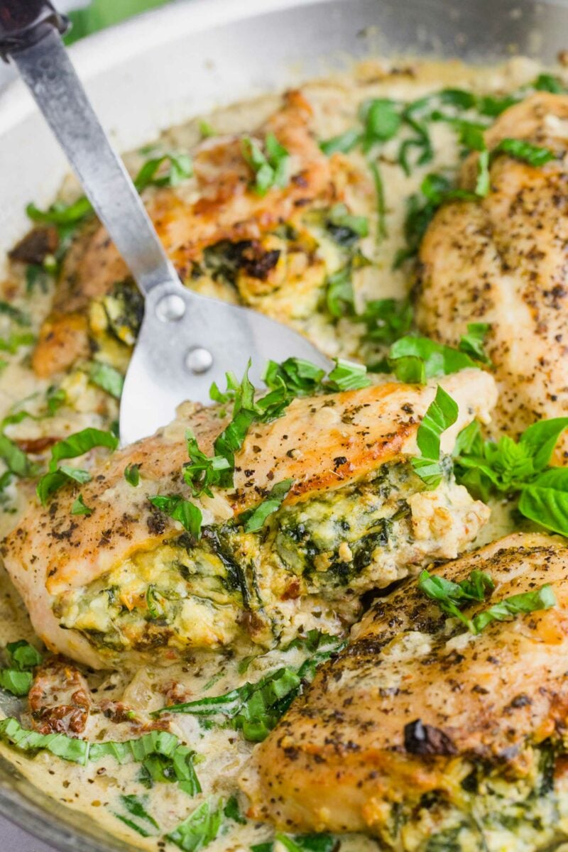 A spatula is lifting a stuffed chicken breast garnished with basil from the skillet.